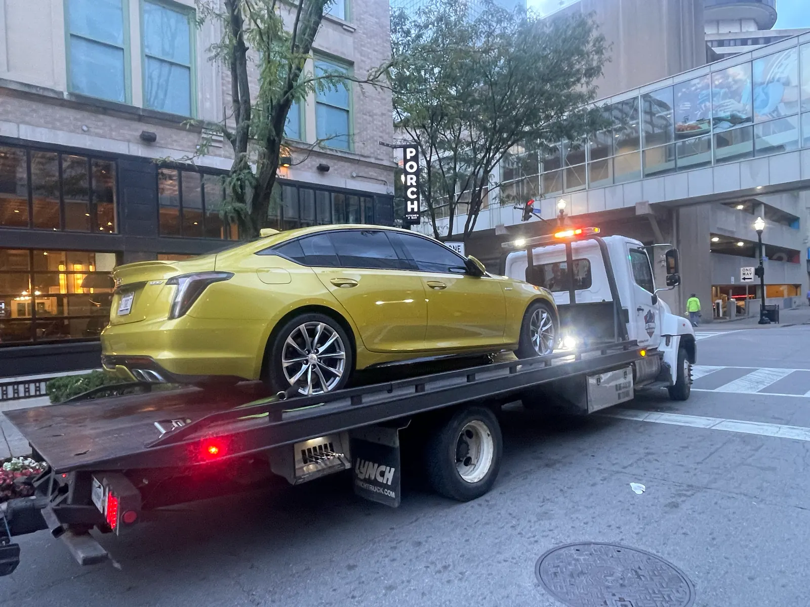 Joe Towing flatbed carrying a yellow sedan on a city street at dusk.