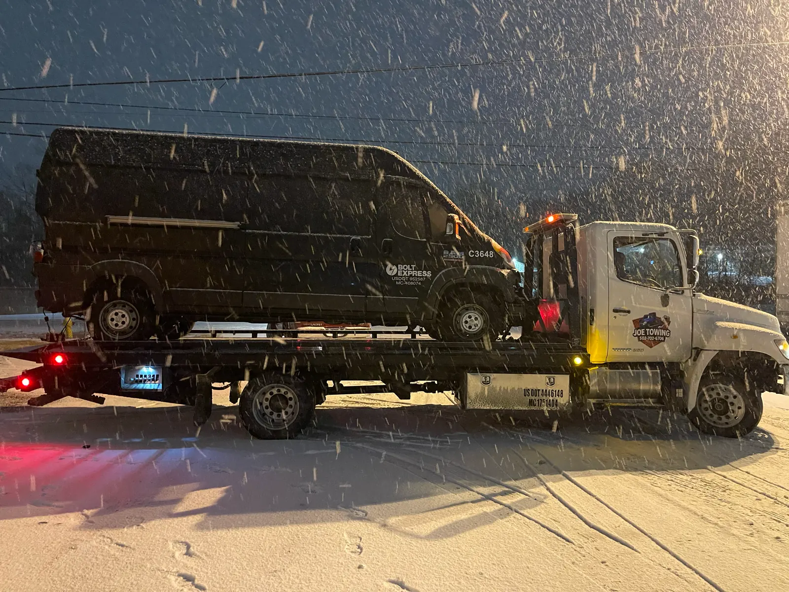 Joe Towing flatbed carrying a black van at night while snow falls.