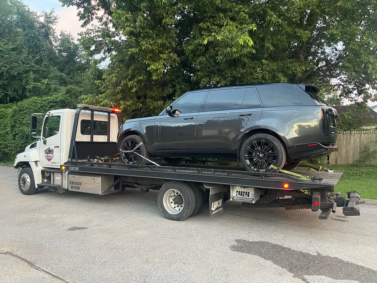Joe Towing flatbed carrying a grey SUV beside a row of trees.